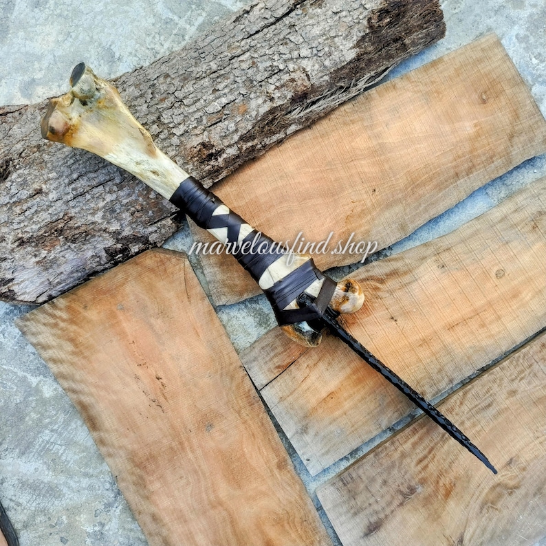 May include: A handcrafted bone dagger with a black leather-wrapped handle and a braided leather blade. The bone has natural markings and is secured with black leather straps. The text "marvelousfind shop" is visible.