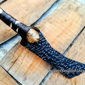 May include: A handcrafted axe with a black, textured blade and a wooden handle wrapped in black leather with white accents. The axe head is a light brown color. The axe is displayed on a wooden surface.