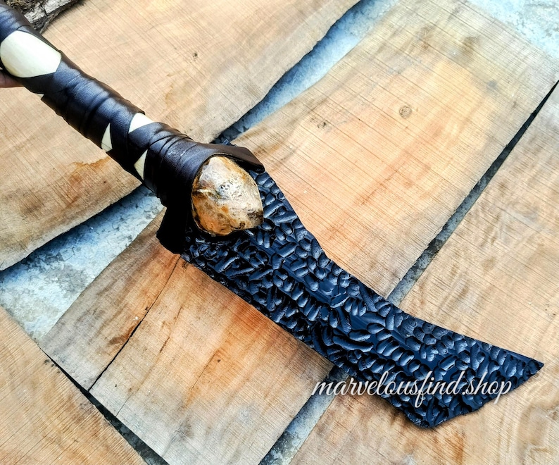 May include: A handcrafted axe with a black, textured blade and a wooden handle wrapped in black leather with white accents. The axe head is a light brown color. The axe is displayed on a wooden surface.