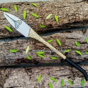 May include: A decorative knife with a silver blade and a gold-coloured handle featuring an ornate design. The handle transitions to a black wrapped grip. The text "marvelousfind shop" is visible on the wooden surface.