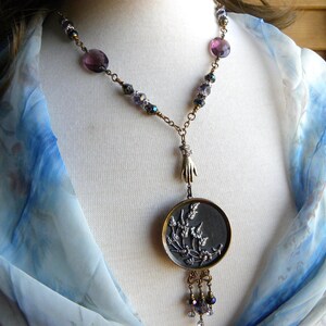May include: A necklace featuring a round pendant with a silver-toned floral relief, suspended from a brass-toned chain. The chain includes purple faceted beads and a small hand-shaped charm. The design incorporates a mix of metallic finishes.