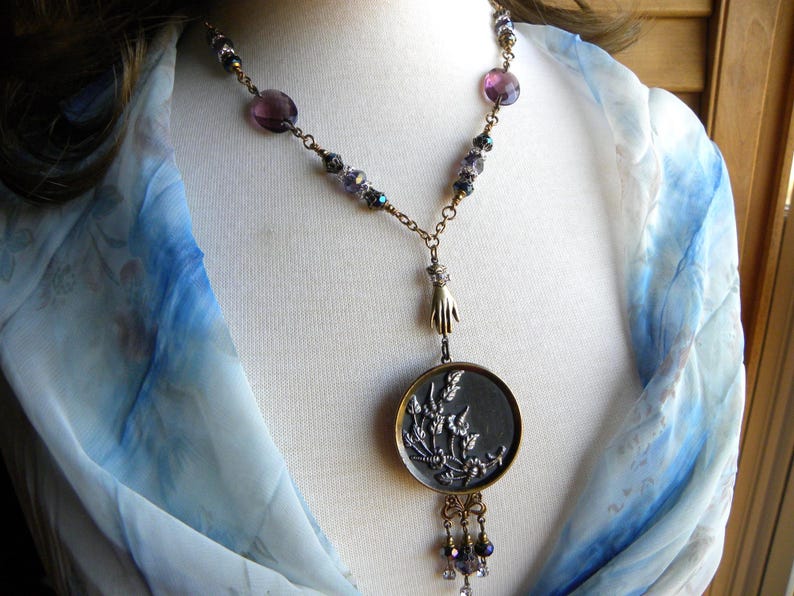 May include: A necklace featuring a round pendant with a silver-toned floral relief, suspended from a brass-toned chain. The chain includes purple faceted beads and a small hand-shaped charm. The design incorporates a mix of metallic finishes.