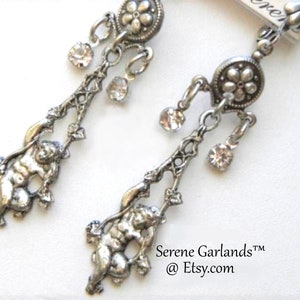May include: Silver dangle earrings with a cherub design, a flower charm, and clear crystal accents. The earrings are labeled "Serene Garlands™ @ Etsy.com".