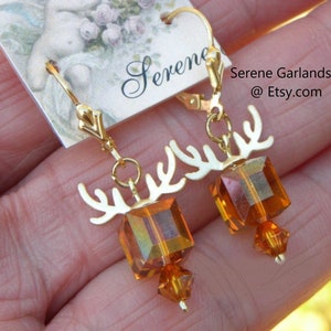 May include: A pair of gold-tone leverback earrings featuring a metallic antler-shaped charm suspended above a square amber-colored crystal bead and a smaller faceted amber crystal drop. The display card includes the text Serene Garlands at Etsy.com.