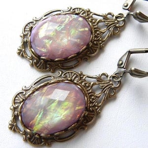 May include: A pair of earrings featuring oval-shaped, faceted pink iridescent stones set in ornate, antique bronze-toned metal frames with lever-back closures. The stones display a shimmering, multi-coloured play of light across their surfaces.