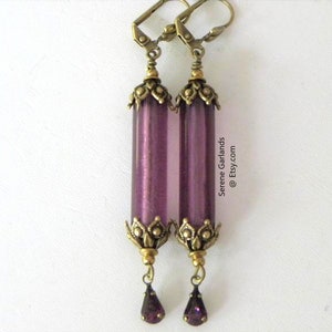 May include: A pair of long, purple earrings with a vintage aesthetic. Each earring features a cylindrical, translucent purple bead, accented with ornate, antique gold-colored metalwork at the top and bottom. A small, purple teardrop-shaped gem hangs from the bottom.