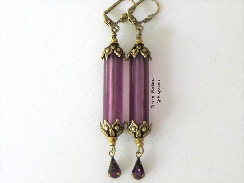 May include: A pair of long, purple earrings with a vintage aesthetic. Each earring features a cylindrical, translucent purple bead, accented with ornate, antique gold-colored metalwork at the top and bottom. A small, purple teardrop-shaped gem hangs from the bottom.
