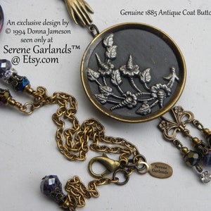 May include: A necklace featuring a round, dark-toned antique coat button from 1885 with a silver-toned floral relief, suspended from a brass chain. The piece includes purple faceted beads, clear crystals, and a small brass hand charm.