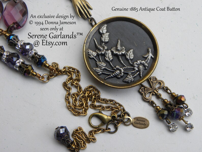May include: A necklace featuring a round, dark-toned antique coat button from 1885 with a silver-toned floral relief, suspended from a brass chain. The piece includes purple faceted beads, clear crystals, and a small brass hand charm.
