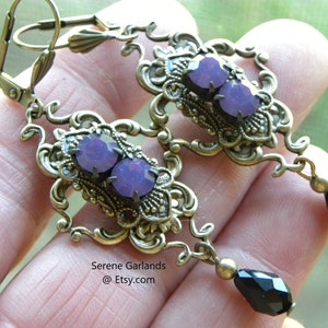 May include: A pair of dangle earrings with a gold-tone filigree design. The earrings feature two purple rhinestones and a black teardrop-shaped bead.