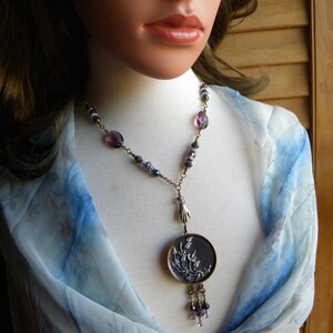 May include: A necklace featuring a round pendant with a raised floral relief design, suspended from a chain adorned with purple and dark faceted beads. A small metallic hand charm connects the pendant to the chain, which ends in a beaded tassel.