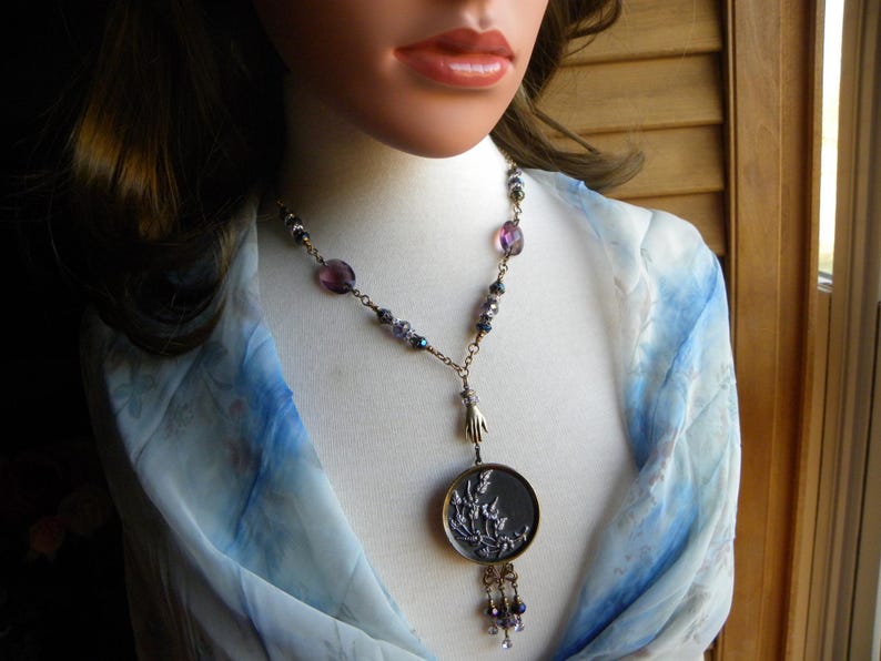 May include: A necklace featuring a round pendant with a raised floral relief design, suspended from a chain adorned with purple and dark faceted beads. A small metallic hand charm connects the pendant to the chain, which ends in a beaded tassel.