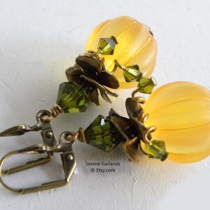 May include: A pair of gold tone earrings with a yellow pumpkin-shaped bead and green crystal accents. The earrings have a hook closure.