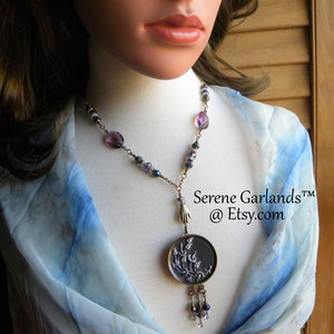 May include: A necklace featuring a round dark metal pendant with a floral relief design, suspended from a chain with purple and blue beads. A small metal hand charm connects the pendant to the chain, which includes dangling beads at the bottom. Text reads Serene Garlands at Etsy.com.
