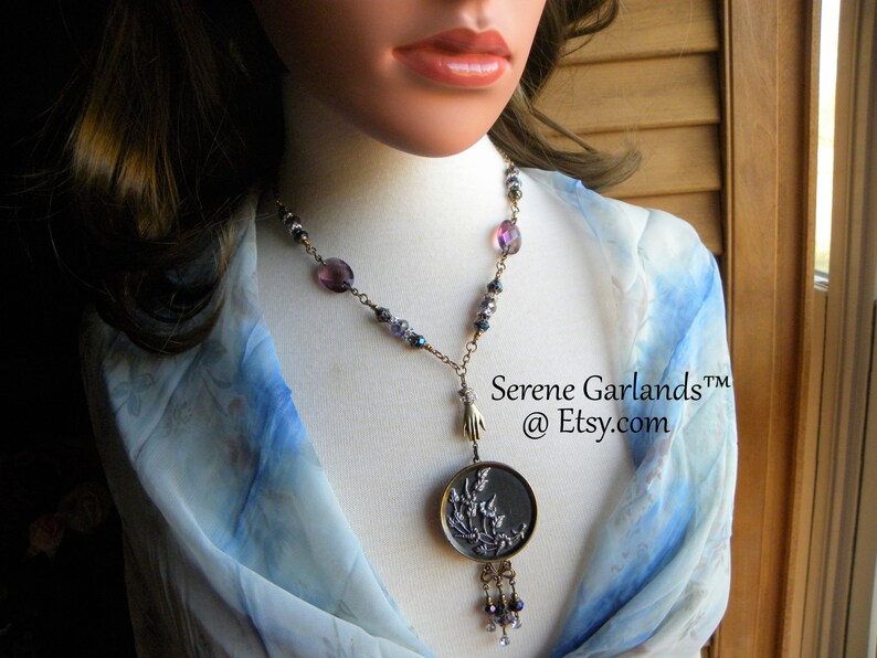 May include: A necklace featuring a round dark metal pendant with a floral relief design, suspended from a chain with purple and blue beads. A small metal hand charm connects the pendant to the chain, which includes dangling beads at the bottom. Text reads Serene Garlands at Etsy.com.