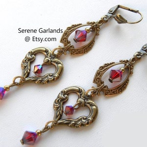 May include: A pair of gold-tone earrings with a heart-shaped design and dangling red crystals. The earrings have a filigree pattern and are accented with small gold beads.