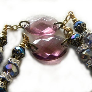 May include: A necklace featuring two large, oval, faceted purple glass beads connected by brass links. The design includes smaller, iridescent blue and purple faceted beads, clear rhinestone spacers, and ornate brass bead caps on a brass chain.