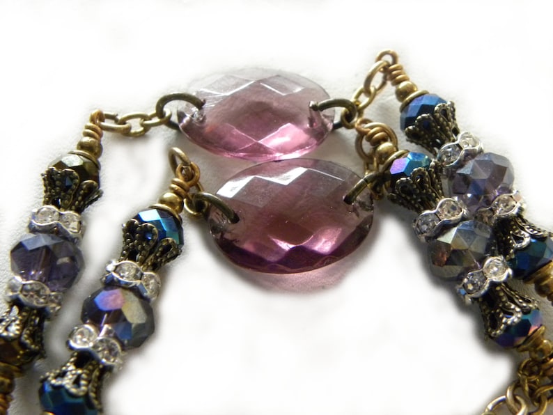 May include: A necklace featuring two large, oval, faceted purple glass beads connected by brass links. The design includes smaller, iridescent blue and purple faceted beads, clear rhinestone spacers, and ornate brass bead caps on a brass chain.