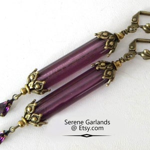 May include: A pair of dangle earrings featuring long, cylindrical purple beads with antique brass-colored accents. Each earring has a decorative floral design at the top and a teardrop-shaped purple crystal at the bottom. The earrings are displayed on a white background.