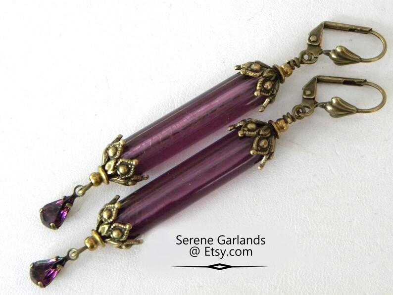 May include: A pair of dangle earrings featuring long, cylindrical purple beads with antique brass-colored accents. Each earring has a decorative floral design at the top and a teardrop-shaped purple crystal at the bottom. The earrings are displayed on a white background.