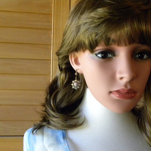 Pode incluir: Uma mulher com cabelo castanho comprido vestindo uma camisa azul claro e um par de brincos de ouro com um design de flor branca.
