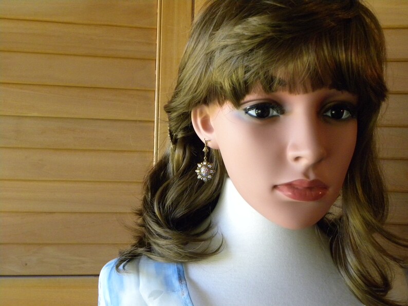 Pode incluir: Uma mulher com cabelo castanho comprido vestindo uma camisa azul claro e um par de brincos de ouro com um design de flor branca.