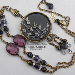 May include: A necklace featuring a round antique button with a silver floral relief on a black background, set in a brass frame. The chain includes purple oval beads, dark faceted beads with clear crystals, a brass hand charm, and a butterfly charm.