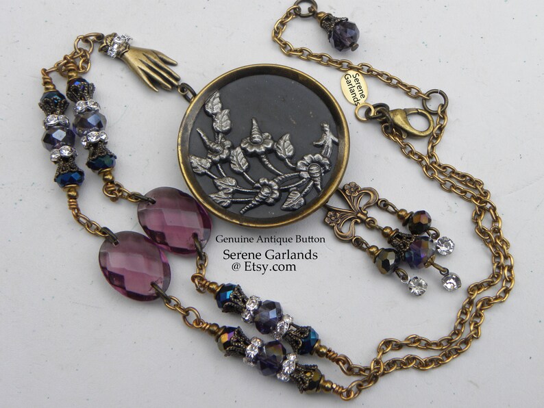 May include: A necklace featuring a round antique button with a silver floral relief on a black background, set in a brass frame. The chain includes purple oval beads, dark faceted beads with clear crystals, a brass hand charm, and a butterfly charm.