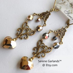 May include: Gold tone dangle earrings with a heart-shaped crystal and a filigree design. The earrings are adorned with clear and iridescent crystals. The earrings are labeled "Serene Garlands™ @ Etsy.com".