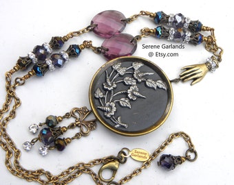 May include: A necklace featuring a central round black button with a silver floral relief, suspended on a brass chain. The design includes purple faceted beads, clear crystals, and a small brass hand charm. Text reads: Serene Garlands @ Etsy.com.