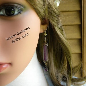 May include: A long, dangling earring with a cylindrical, purple and clear glass bead design. The earring has ornate gold-colored metal accents and a small purple bead at the bottom. The text "Serene Garlands @ Etsy.com" is visible.