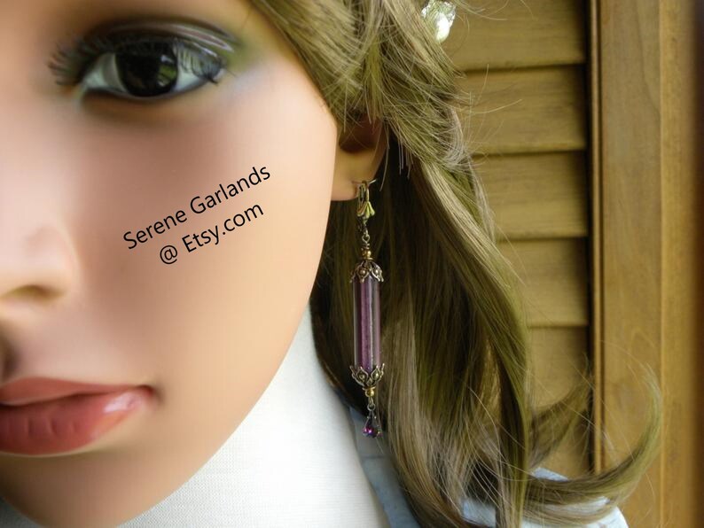 May include: A long, dangling earring with a cylindrical, purple and clear glass bead design. The earring has ornate gold-colored metal accents and a small purple bead at the bottom. The text "Serene Garlands @ Etsy.com" is visible.