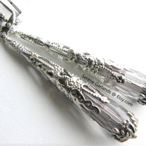 May include: Silver dangle earrings with a clear, faceted crystal drop and intricate filigree details. The earrings are approximately 2 inches long.