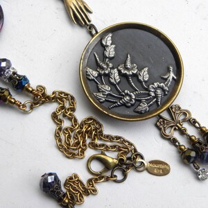 May include: A necklace featuring a round brass pendant with a black inlay and a silver-toned floral relief. The chain includes purple faceted beads, clear crystals, and a small brass hand charm. A tag on the chain is engraved with the text Mourning Era.
