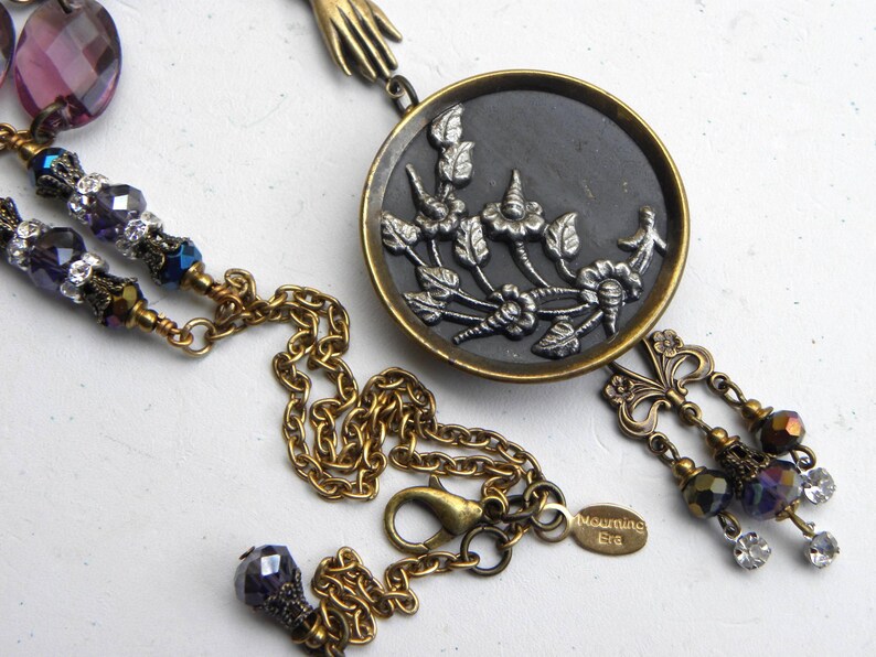 May include: A necklace featuring a round brass pendant with a black inlay and a silver-toned floral relief. The chain includes purple faceted beads, clear crystals, and a small brass hand charm. A tag on the chain is engraved with the text Mourning Era.
