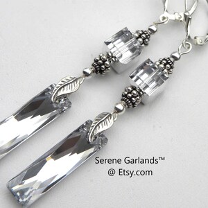 May include: A pair of drop earrings featuring clear rectangular crystal pendants, silver-toned leaf charms, and small clear cube beads. The earrings are finished with silver-toned metal findings and lever-back closures. Text reads Serene Garlands at Etsy.com.