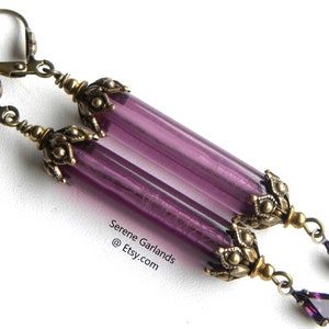 May include: A pair of long, dangling earrings with purple glass tube beads and antique brass-colored metal accents. Each earring features a teardrop-shaped purple crystal at the bottom. The text "Serene Garlands @ Etsy.com" is visible.