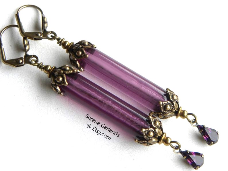 May include: A pair of long, dangling earrings with purple glass tube beads and antique brass-colored metal accents. Each earring features a teardrop-shaped purple crystal at the bottom. The text "Serene Garlands @ Etsy.com" is visible.