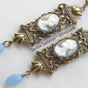 May include: A pair of antique-style earrings. Each earring features a cameo portrait within an ornate, bronze-toned frame. A light blue teardrop-shaped bead dangles from the bottom. The text "Serene Garlands @Etsy.com" is visible.