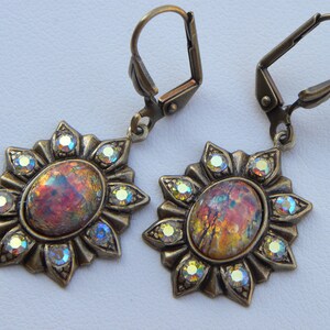 Pode incluir: Um par de brincos dourados com pedras de opala iridescente engastadas em um design de sol. Cada brinco tem um fecho de gancho.