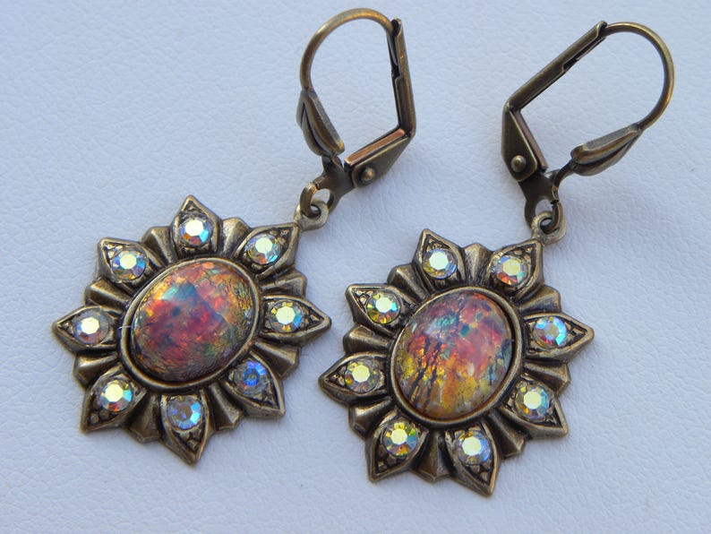Pode incluir: Um par de brincos dourados com pedras de opala iridescente engastadas em um design de sol. Cada brinco tem um fecho de gancho.