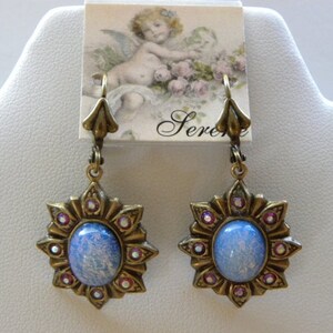 Pode incluir: Um par de brincos pendentes com um caboch&atilde;o de vidro azul iridescente engastado em um design de estrela dourada com strass rosa. Os brincos t&ecirc;m um fecho de gancho.