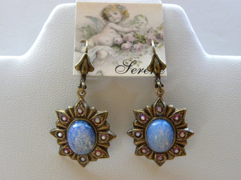 Pode incluir: Um par de brincos pendentes com um caboch&atilde;o de vidro azul iridescente engastado em um design de estrela dourada com strass rosa. Os brincos t&ecirc;m um fecho de gancho.