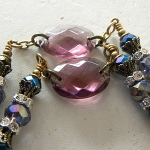 May include: A necklace featuring two large, oval, faceted purple glass beads connected by brass chains. The design includes smaller, iridescent blue and purple faceted beads, clear rhinestone spacers, and ornate brass bead caps throughout.