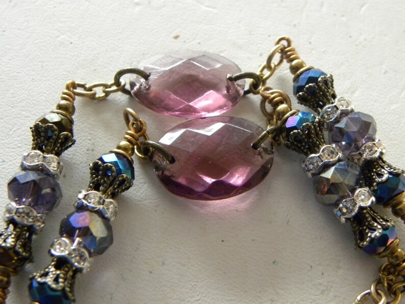 May include: A necklace featuring two large, oval, faceted purple glass beads connected by brass chains. The design includes smaller, iridescent blue and purple faceted beads, clear rhinestone spacers, and ornate brass bead caps throughout.