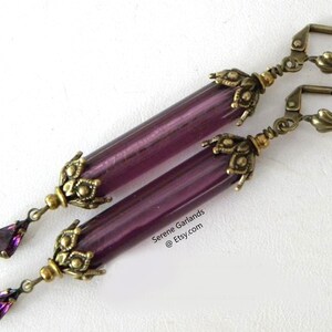 May include: A pair of long, dangle earrings with a vintage aesthetic. Each earring features a translucent, cylindrical purple bead, accented with ornate, antique brass-toned metalwork. A teardrop-shaped purple crystal dangles from the bottom.
