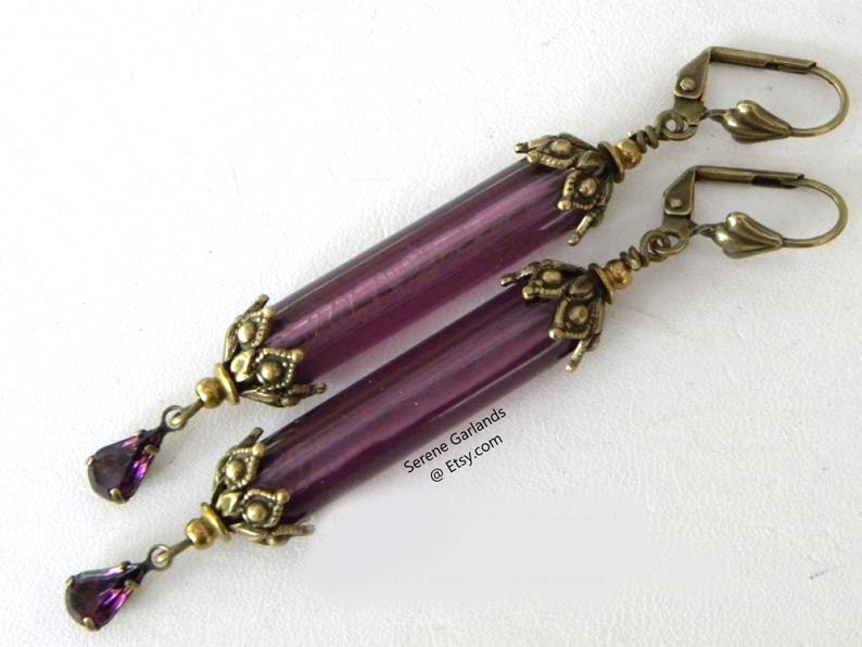 May include: A pair of long, dangle earrings with a vintage aesthetic. Each earring features a translucent, cylindrical purple bead, accented with ornate, antique brass-toned metalwork. A teardrop-shaped purple crystal dangles from the bottom.