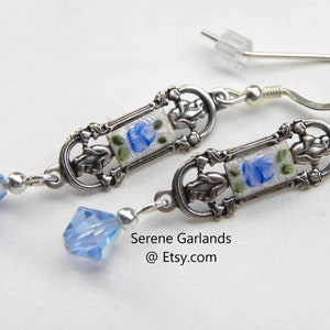 May include: A pair of silver earrings with blue and white enamel floral designs and blue crystal accents. The earrings are dangle style and have a hook closure.  Serene Garlands @Etsy.com