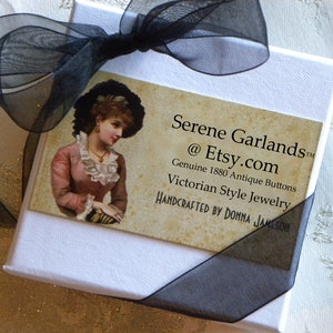 May include: A white square gift box tied with a black sheer ribbon. A rectangular card is attached, featuring an illustration of a person in 19th-century attire and the text: Serene Garlands @ Etsy.com Genuine 1880 Antique Buttons Victorian Style Jewelry.