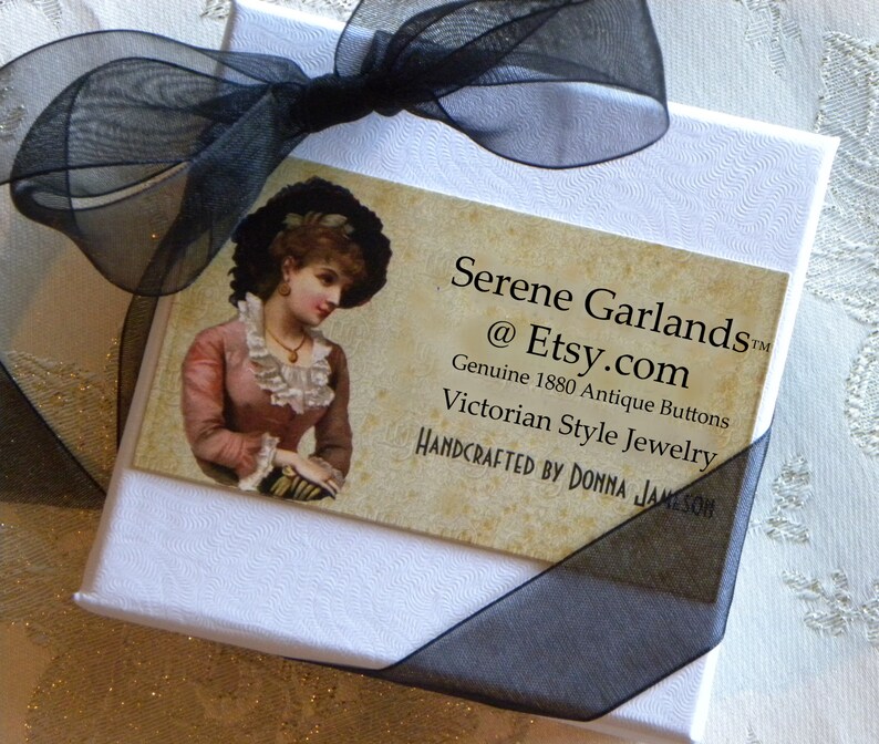 May include: A white square gift box tied with a black sheer ribbon. A rectangular card is attached, featuring an illustration of a person in 19th-century attire and the text: Serene Garlands @ Etsy.com Genuine 1880 Antique Buttons Victorian Style Jewelry.